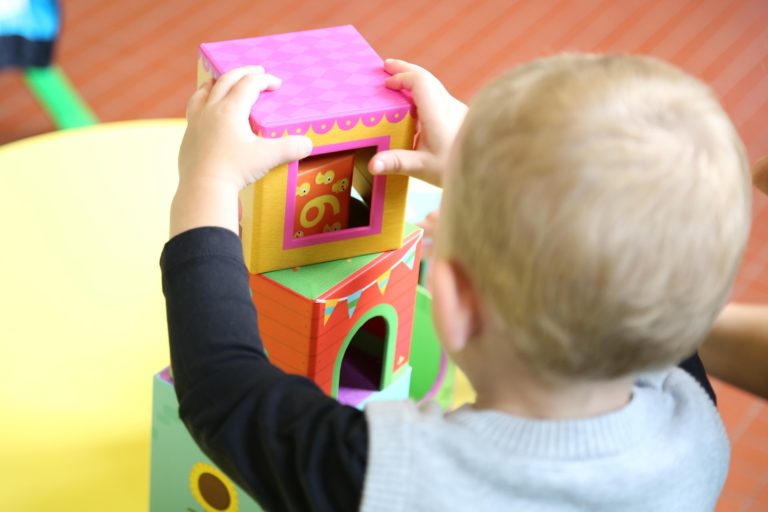 Ett litet barn bygger ett färgglatt torn med lådor på ett bord, fotograferat bakifrån i en förskolemiljö.